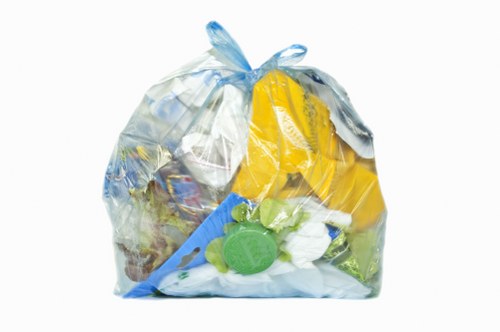 A clear plastic rubbish bag filled with various household waste items, including yellow packaging, white tissues or paper, green plastic bottles, and other miscellaneous refuse, sits on a plain white background. The bag is tightly closed at the top with a tied knot, and its semi-transparent material reveals the mixture of different materials inside. The plastic bag's surface has a crinkled texture, and the waste materials are visible through the thin plastic, indicating it contains general household rubbish. This scene illustrates typical rubbish collected for disposal or removal, relevant to commercial waste services such as those provided by Commercial Waste Mortlake in the Mortlake area, with focus on waste management and rubbish collection in London postcodes.