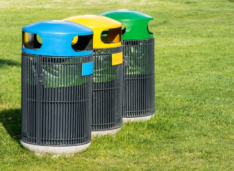 Three outdoor litter bins positioned on a well-maintained grassy area, each with a cylindrical metal body featuring vertical slats and a domed, colourful plastic lid in blue, yellow, and green. The bins are aligned side by side with small rectangular slots on their front panels for waste disposal, and they are set on a paved surface at the edge of a green lawn, suggesting a public or communal outdoor space in Mortlake. The weather appears clear with natural light illuminating the scene, emphasizing the vibrant colours of the lids against the lush grass. These bins are part of waste management services supplied by Commercial Waste Mortlake, suitable for residential or commercial areas around the local postcode and town, supporting rubbish collection and disposal efforts in the area.