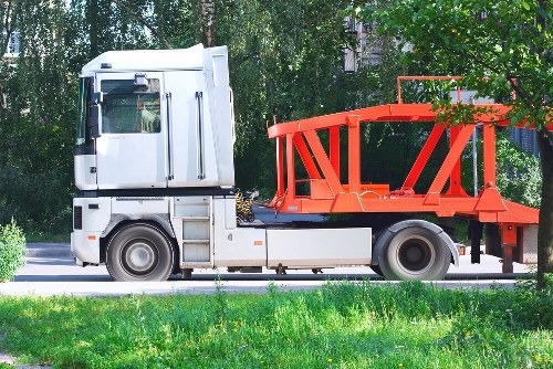 A white commercial truck with a rectangular cabin and black detailing, parked on a paved driveway next to a grassy verge. The truck is equipped with a large red metal loading platform or scrap container on its chassis, featuring vertical support beams and angled bracing. The platform is empty, with no visible waste or debris, and appears to be ready for loading or transport. In the background, dense green foliage and trees provide a natural setting, while a hint of a building structure is partially visible behind the trees. The scene is illuminated by natural daylight, casting minimal shadows, suggesting overcast or diffuse sunlight. The overall environment indicates a location suitable for rubbish collection or waste removal services, such as those provided by Commercial Waste Mortlake, servicing the Mortlake area. The image emphasizes the vehicle’s purpose in rubbish or waste transport, with a practical, professional appearance fitting for commercial waste removal activities in the local area around Richmond upon Thames.