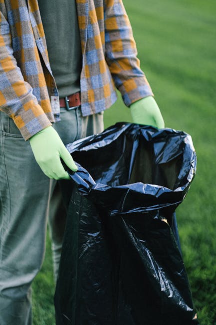 A waste collection worker stands next to a large industrial rubbish disposal vehicle on a street at dusk. The man is wearing a high-visibility jacket with orange and reflective silver stripes, and dar