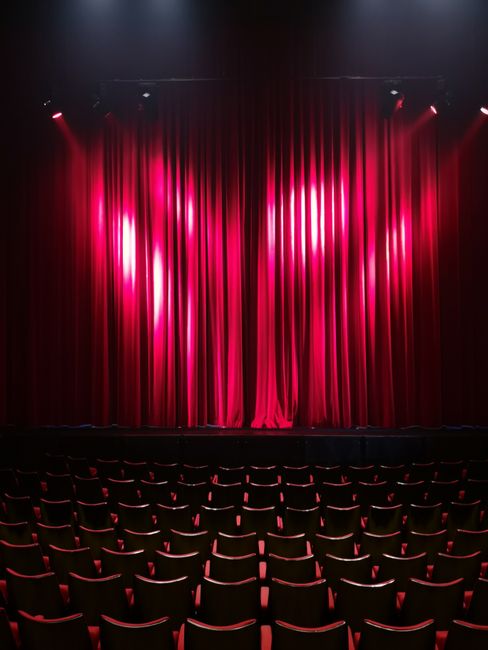 The image depicts a stage with closed red velvet curtains, illuminated softly by overhead spotlights creating a warm glow. In the foreground, multiple rows of black theatre seats with red trim are vis