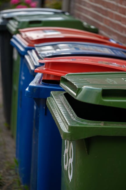 A black wheeled rubbish bin labeled with the name 'St. John’s' is positioned on a paved sidewalk beside a curb in a residential or urban street at night. The bin is filled with a variety of waste ma