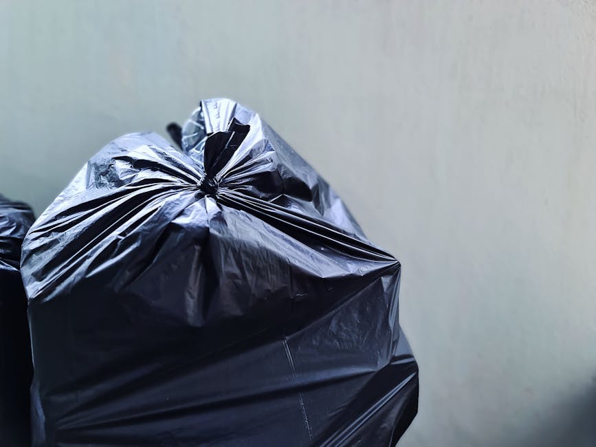 A worker wearing a grey beanie, dark clothing, and a high-visibility vest stands on a modern train station platform beside a red metal trolley overloaded with black, orange, and white garbage bags, as